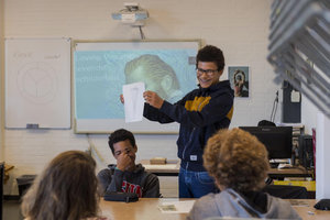 Een jongen met donkere krullen en een bril toont lachend zijn opdracht voor de klas. Op de achtergrond het digibord met een afbeelding uit de film Loving Vincent. Foto: Jan-Kees Steenman